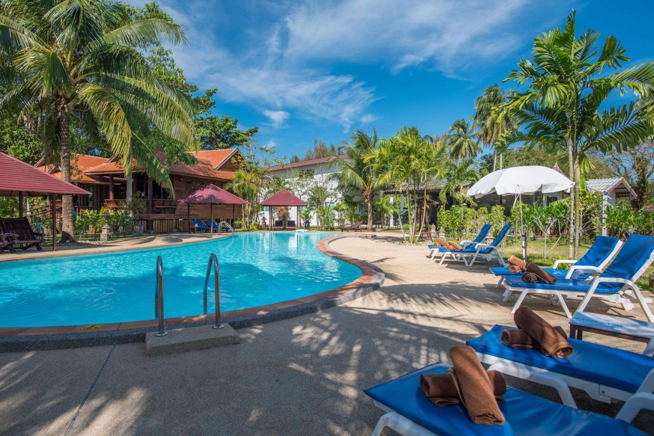 Tranquil swimming pool at ThaiLife Homestay Resort & Spa surrounded by palm trees, traditional Thai villas, and blue lounge chairs with rolled towels