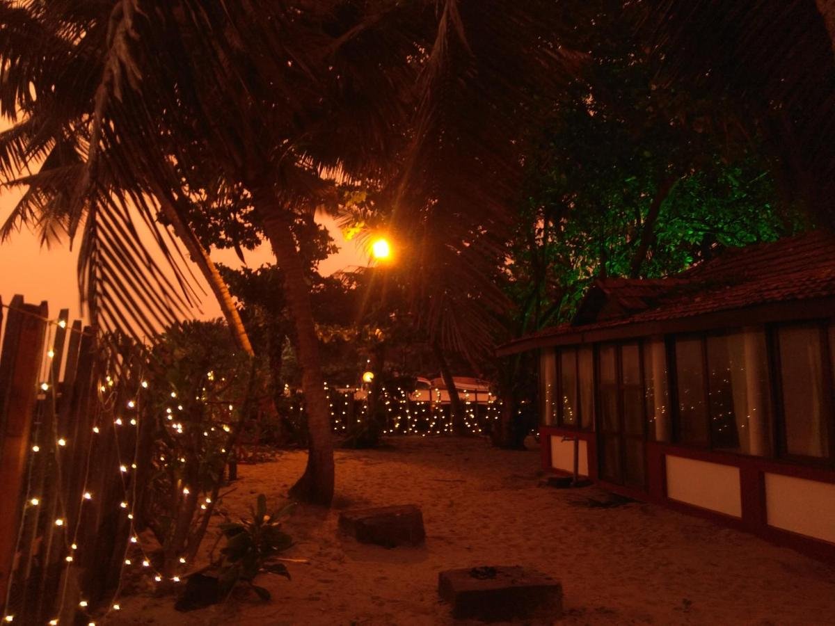Evening atmosphere at Sambodhi India Yoga Retreat with string lights, palm trees, traditional cabins, and sunset over sandy grounds