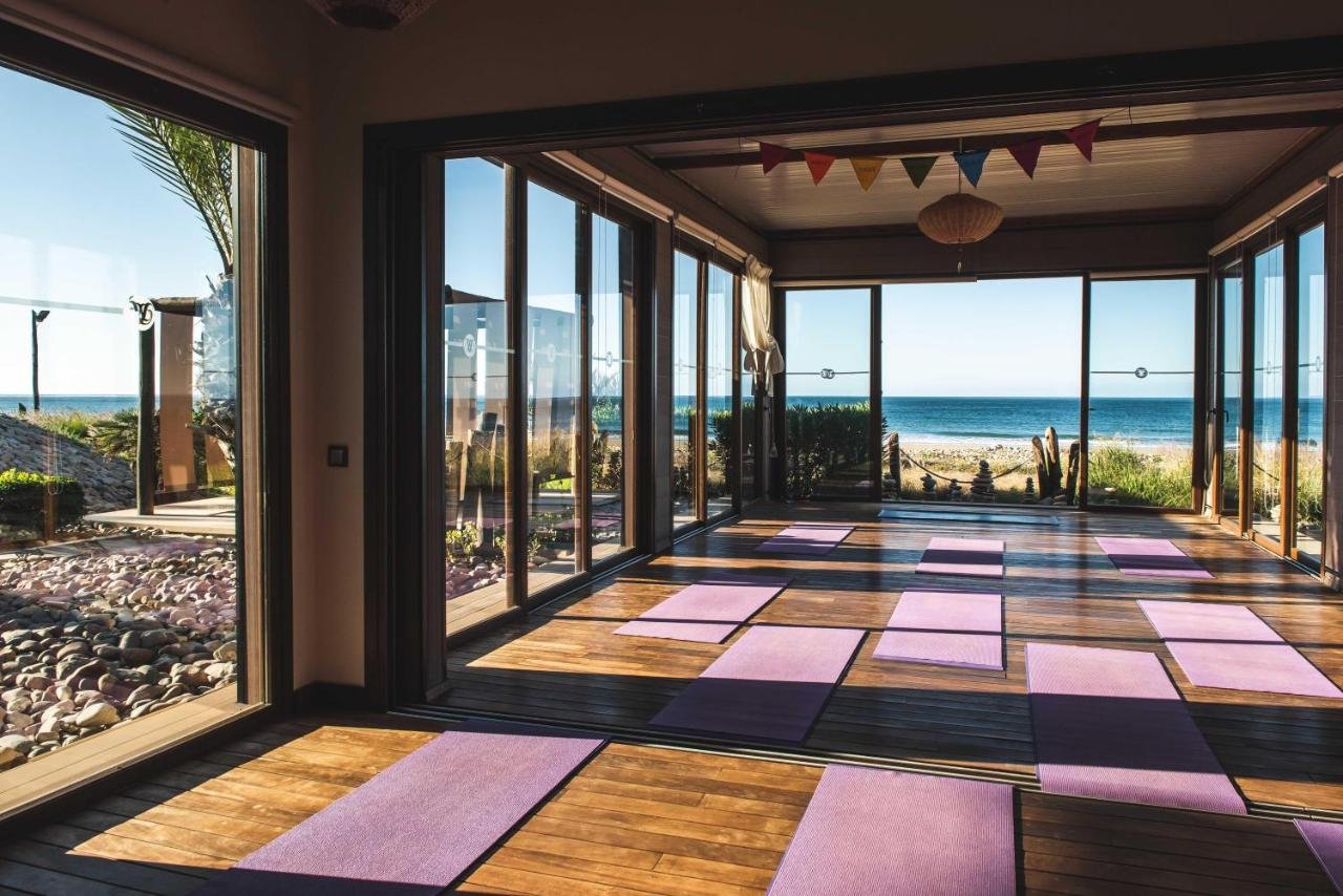 Interior of beachfront yoga studio at Morocco surf yoga retreat with purple yoga mats arranged on wooden floor and panoramic ocean views.